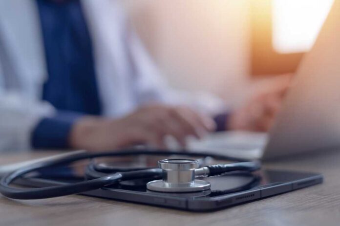 Close-up,Of,Doctor,Working,On,Laptop,Computer,With,Digital,Tablet A stethoscope resting on a tablet with a blurred medical professional in the background typing on a laptop