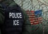 What Happened During Maine ICE Raid? A police officer in a tactical vest stands near a barbed wire fence with an American flag in the background