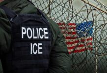 What Happened During Maine ICE Raid? A police officer in a tactical vest stands near a barbed wire fence with an American flag in the background