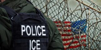 A police officer in a tactical vest stands near a barbed wire fence with an American flag in the background