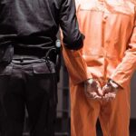 Firefighters Catch Alleged Park Predator Prisoner in orange jumpsuit handcuffed with guard standing beside
