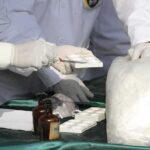 Scientists in lab coats handling white powder sample with tools