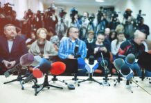 Row of microphones at a press conference with audience and cameras behind