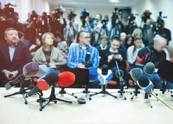 shutterstock_1244384347 Row of microphones at a press conference with audience and cameras behind