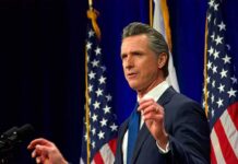 Man in suit speaking at podium with U.S. flags behind him