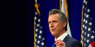 Man in suit speaking at podium with U.S. flags behind him