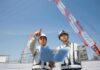 Two construction workers reviewing plans at a site with cranes