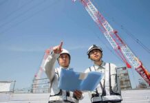 Two construction workers reviewing plans at a site with cranes