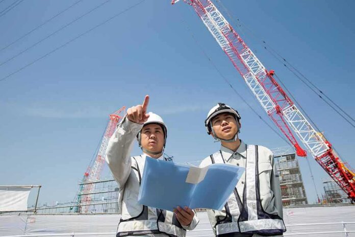 shutterstock_2154192201 Two construction workers reviewing plans at a site with cranes