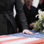 Mourner rests hand on flag-draped casket with flower arrangement