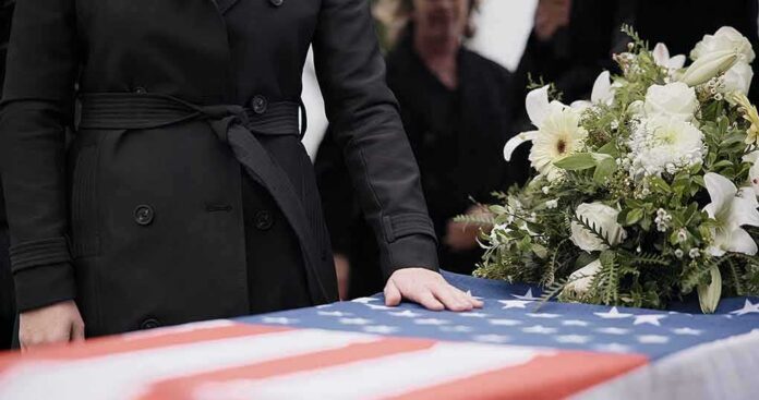 Mourner rests hand on flag-draped casket with flower arrangement
