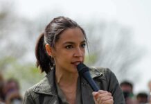 Affluent Suburb Twist In AOC Story AOC speaking into a handheld microphone outdoors