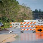 Isaias Roars In – Evacuations Loom Road closed barricades blocking a flooded rural highway with detour sign
