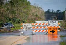 Isaias Roars In – Evacuations Loom Road closed barricades blocking a flooded rural highway with detour sign